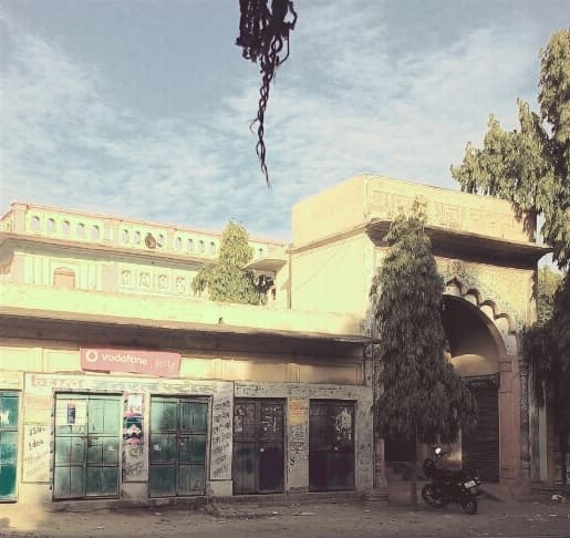 कोइरीपुर का शिवाला मंदिर, उसके आगे बंद शटरों की क़तार और हिंदी में लिखा साइनबोर्ड। किसी खुले संग्रह में मिला यह कोइरीपुर का पहला और अब तक का एकमात्र सड़क-स्तरीय छायाचित्र है। रिज़ॉल्यूशन कम है और उस पर फ़िल्टर लगा है — इसे बदला नहीं गया, क्योंकि अभिलेख वस्तु को वैसे ही रखता है जैसी वह मिली।